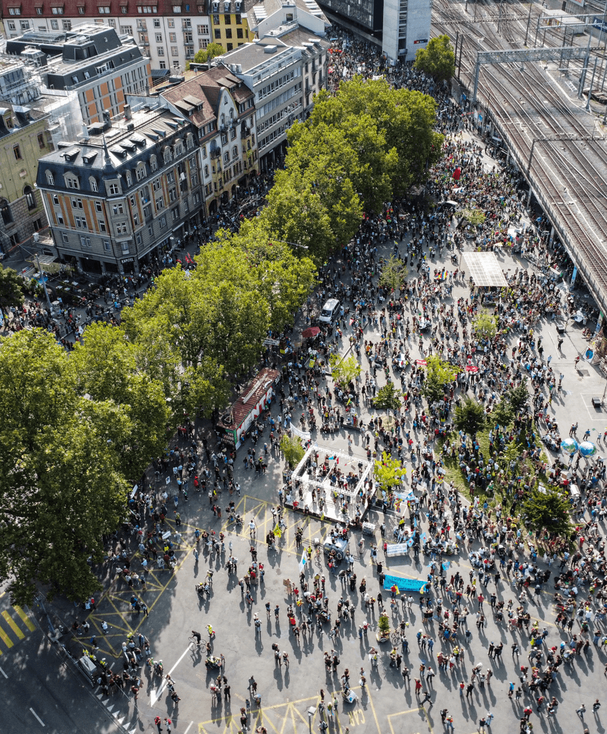 Eine Stadt aus der Vogelperspektive. Es versammeln sich viele Leute unter Bäumen und um Häuser um eine Bühne._FR